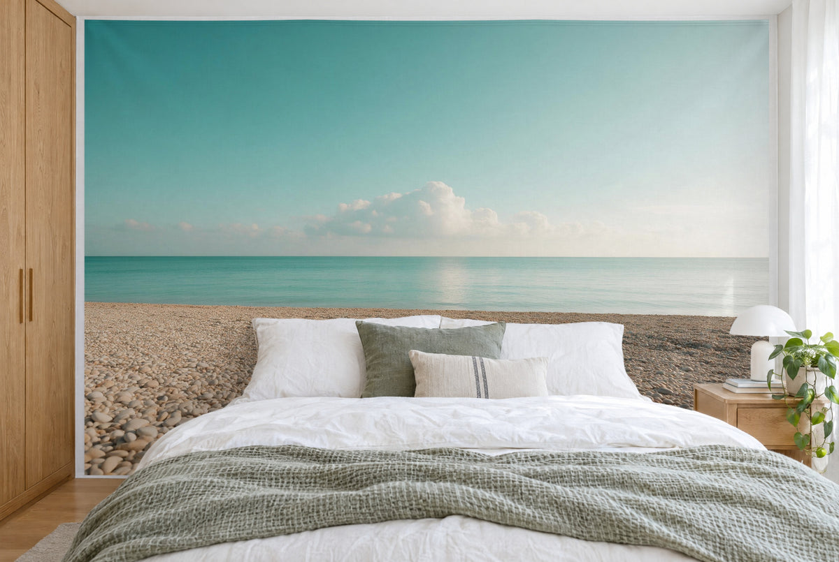 Bedruckter Stoff im XXL-Format: Kieselstrand und türkises Wasser unter Wolken, wandfüllend hinter dem Bett