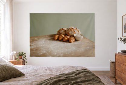 Wandtuch Bauernbrot und Croissants im hellen Schlafzimmer gegenüber dem Bett, rechts eine Holzkommode – Braun und Salbeigrün