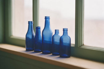 Botellas de vidrio azul en fila sobre un alféizar claro ante un marco verde, tapiz impreso en tela – azul y beige