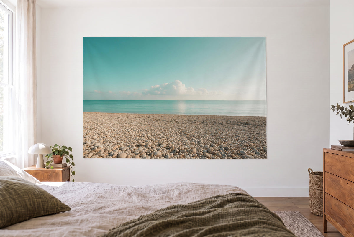 Wandtuch Kieselstrand unter Wolkenhimmel im hellen Schlafzimmer gegenüber dem Bett, rechts eine Holzkommode – Beige und Blau