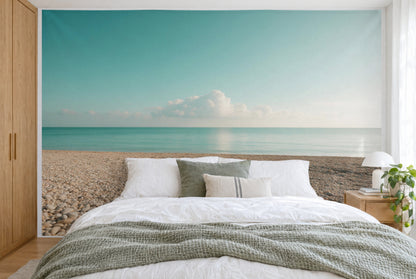 Bedruckter Stoff im XXL-Format: Kieselstrand und türkises Wasser unter Wolken, wandfüllend hinter dem Bett