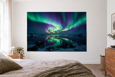 Tapiz auroras boreales lago polar en dormitorio claro frente a la cama, cómoda de madera a la derecha – verde y violeta