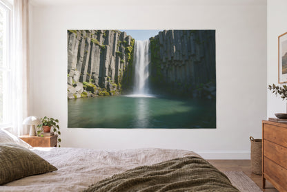 Tapiz cascada sobre rocas de basalto en un dormitorio claro frente a la cama, cómoda de madera a la derecha – gris y verde
